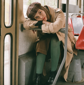 Tall young girl sitting down. She's wearing a hat, a long brown dress, a coat, green tights and black boots. Her name is Ekaterina. Learn Spanish through short stories.